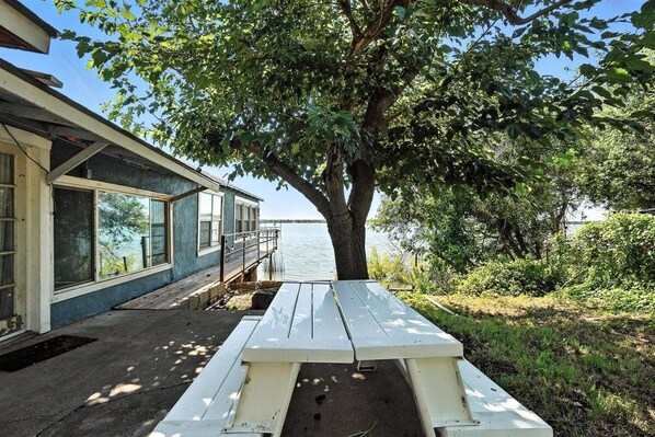 Un chalet au bord du lac, avec un coin repas aménagé sous un arbre, offrant une vue sur l’eau.