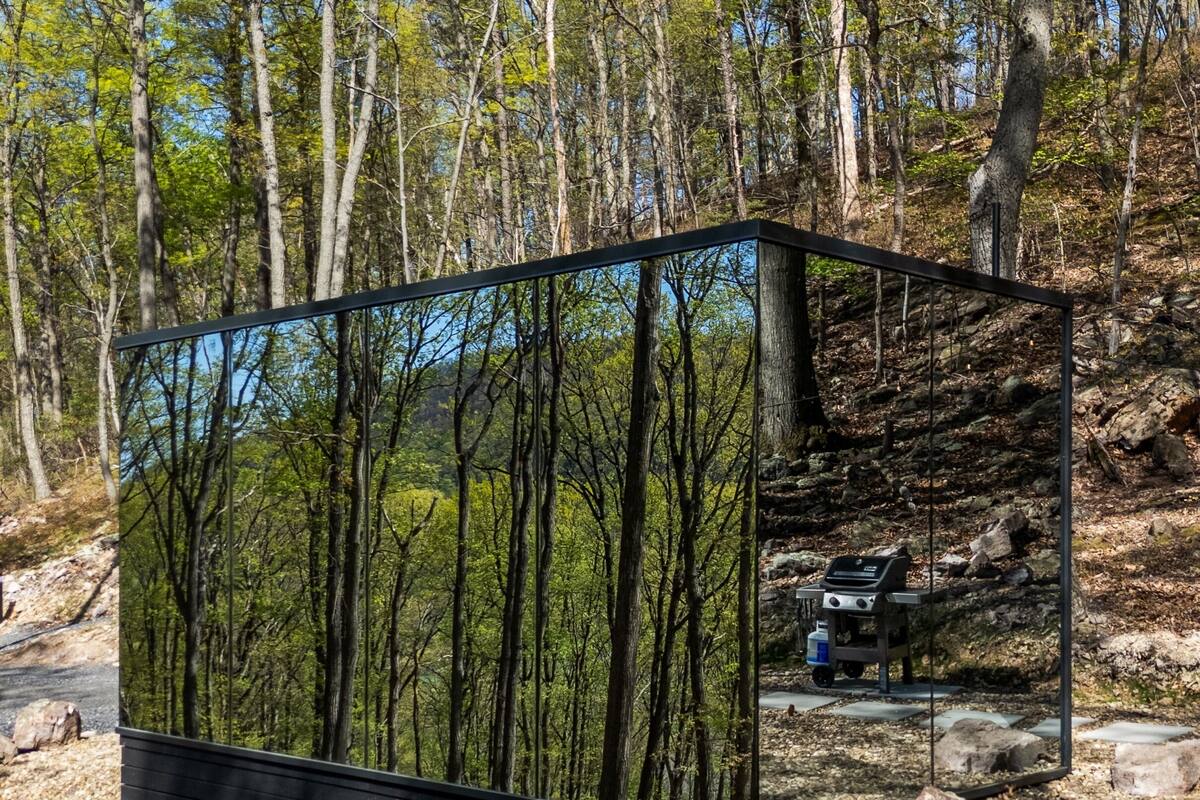 Una estructura moderna y rectangular con fachada espejada que refleja un paisaje boscoso. En su interior, hay una pequeña kitchenette con estufa y dispensador de agua.