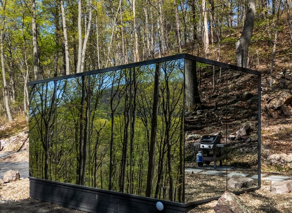 A modern, rectangular structure with a mirrored exterior reflecting a forested landscape. Inside, there is a small kitchenette with a stove and a water dispenser. - Mirror Villas - Modern Retreat with private hot tubs and mountain view (Mercersburg)