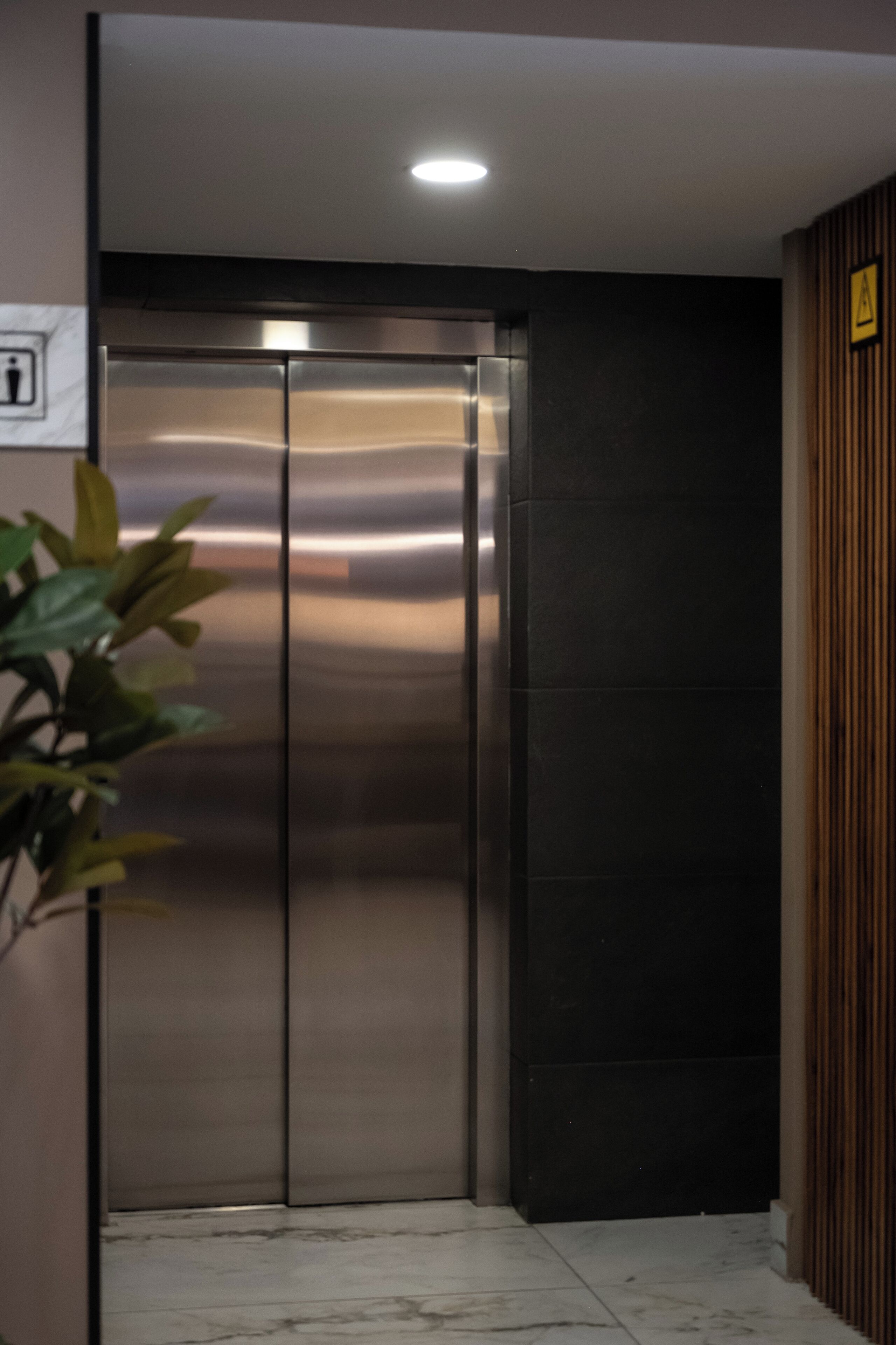 Un elevador moderno con detalles metálicos, piso de mármol y una planta en su izquierda.