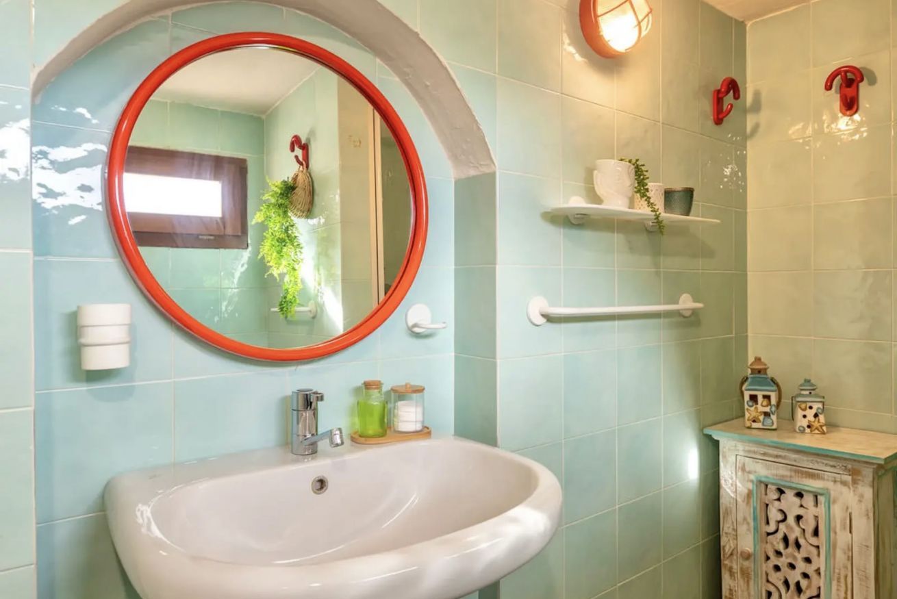 A bathroom with a white sink, a round mirror with a red frame, and light blue tiled walls.
