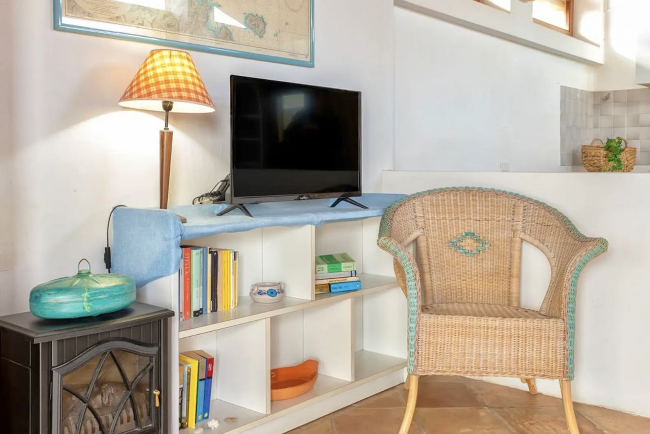Una sala de estar con televisión, una silla de mimbre, una estantería y una lámpara.