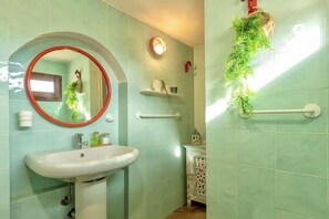 Un baño con un lavamanos blanco, un espejo redondo con marco rojo y azulejos verde claro. Se ven estantes con artículos de tocador y una planta en la pared.
