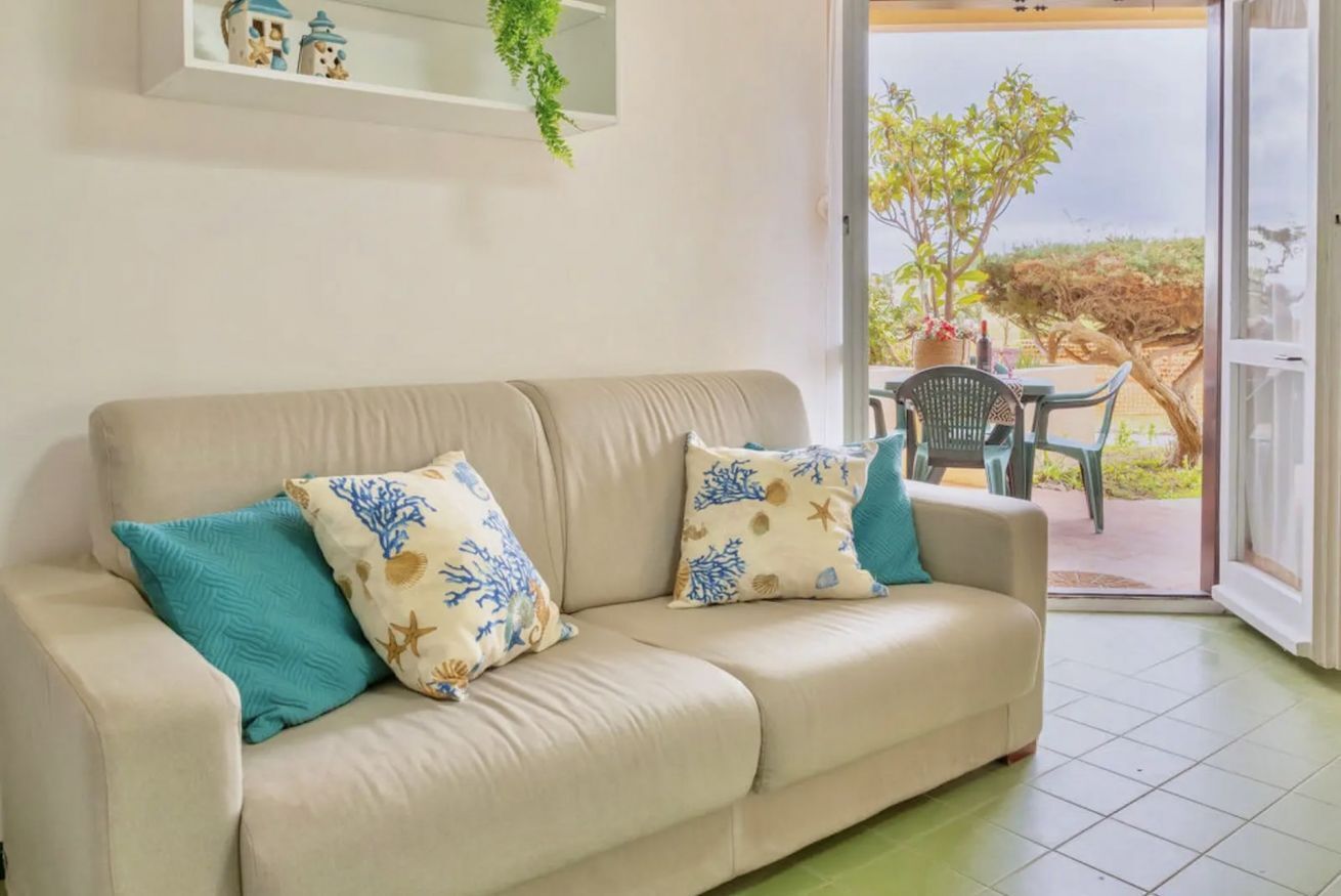 Een beige bank met twee decoratieve kussens, een klein schapje met versieringen, en uitzicht op een terras met een tafel en stoelen.