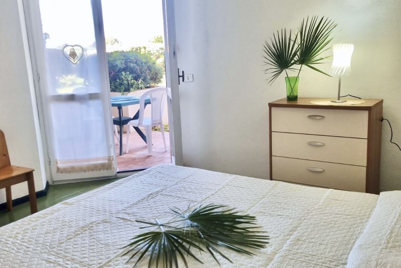 A bedroom with a bed, a nightstand with a lamp, a chair, and a view through a glass door to an outdoor patio.