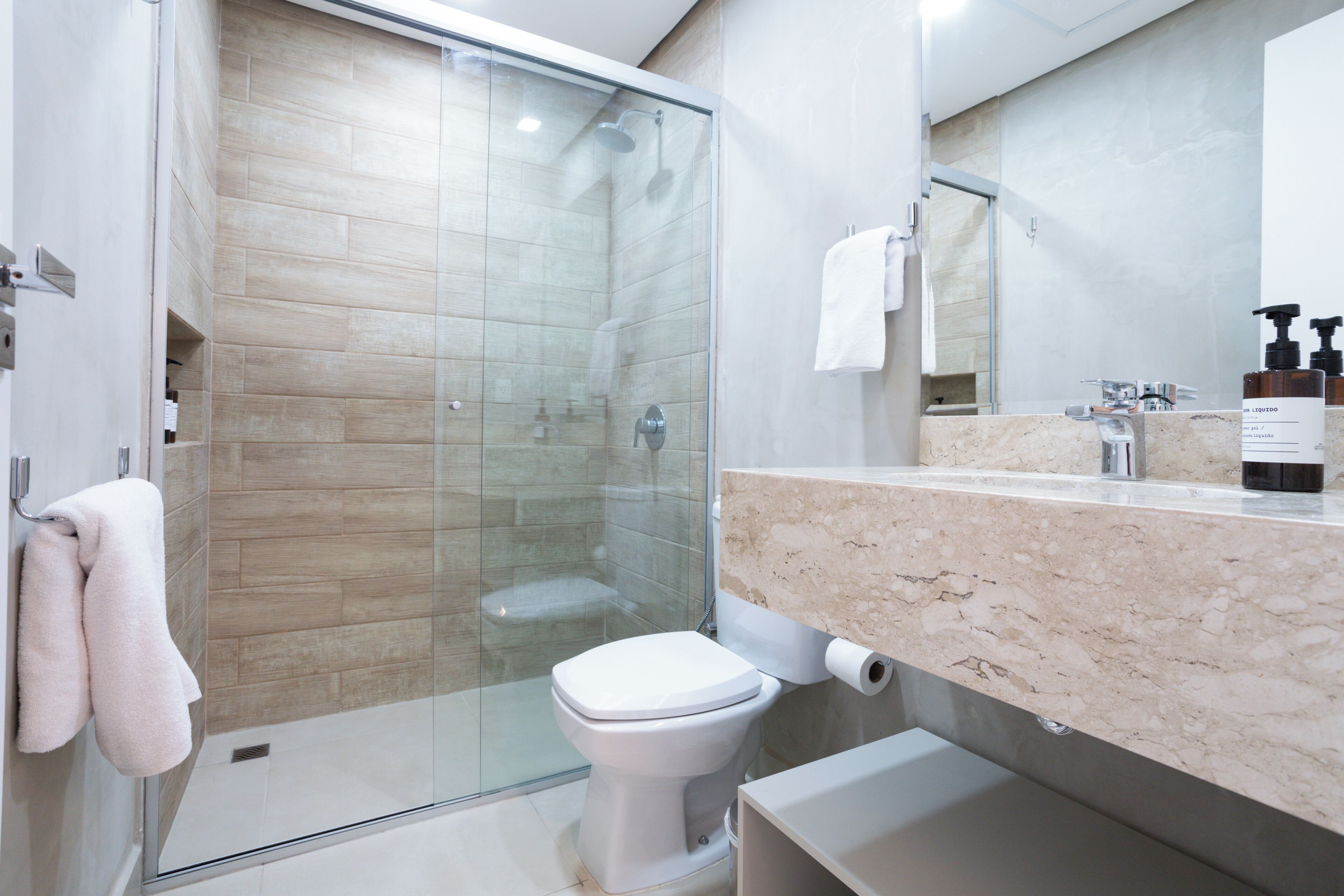 Un baño moderno con ducha envuelta en vidrio, un inodoro blanco y una encimera de mármol con artículos de aseo.