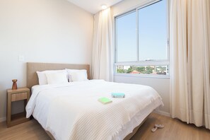 A neatly made bed with a white comforter and pillows, a bedside table with a lamp, and a window with curtains. - Sky Stays at Life Mariscal (Asunción)
