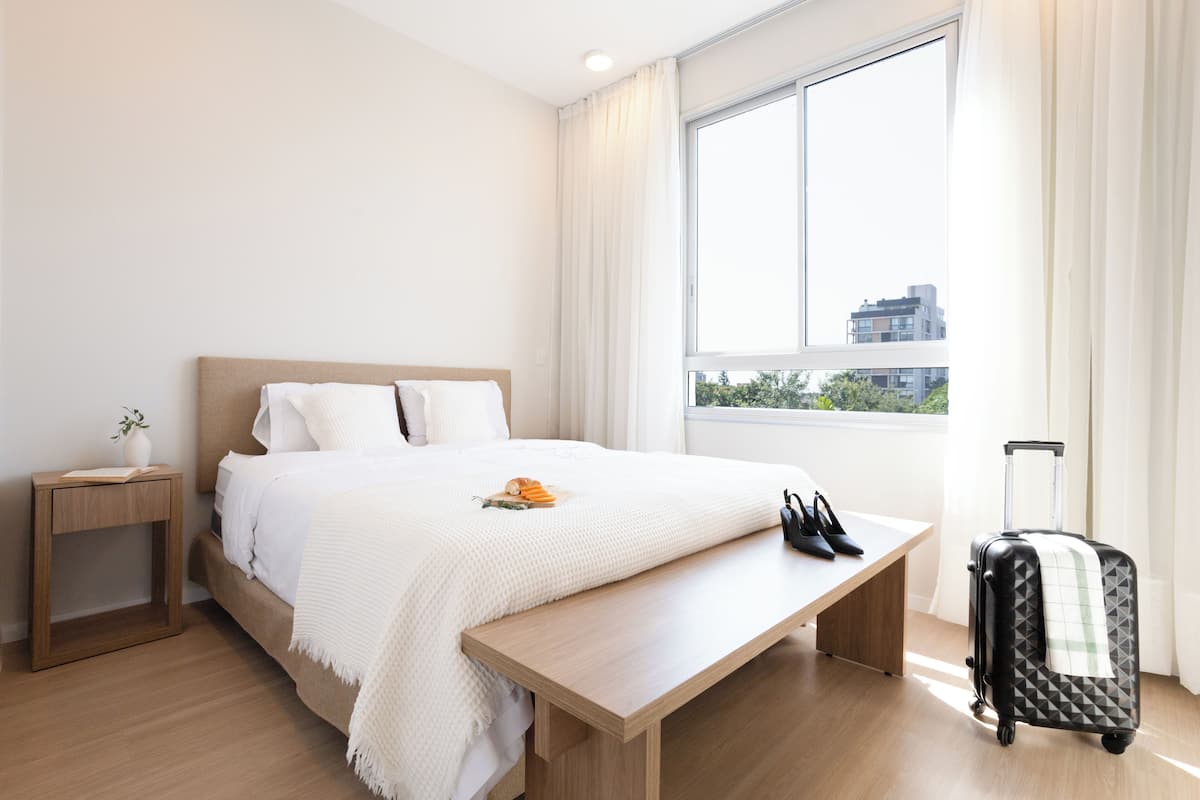 Una camera d'albergo con un letto, un comodino, una panca, una valigia e una finestra con tende.