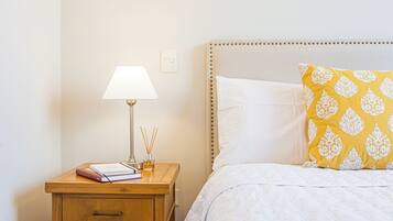 Una cama bien tendida con un cojín estampado, una lámpara de noche y una mesita de noche con libros y una lámpara.