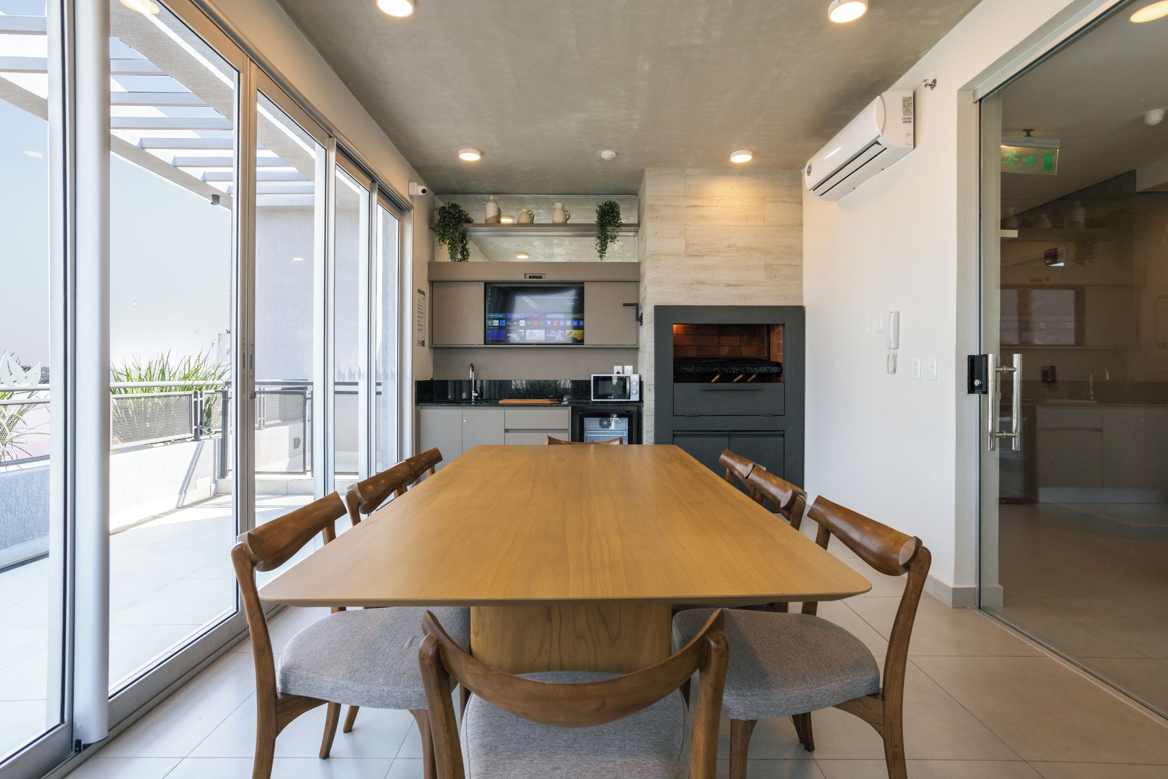 A modern dining area with a large wooden table and cushioned chairs, a kitchen in the background, and large windows offering a view of the outdoors.