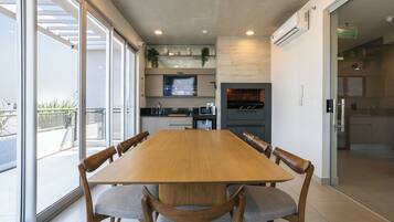 A modern dining area with a large wooden table and cushioned chairs, a kitchen in the background, and large windows offering a view of the outdoors.