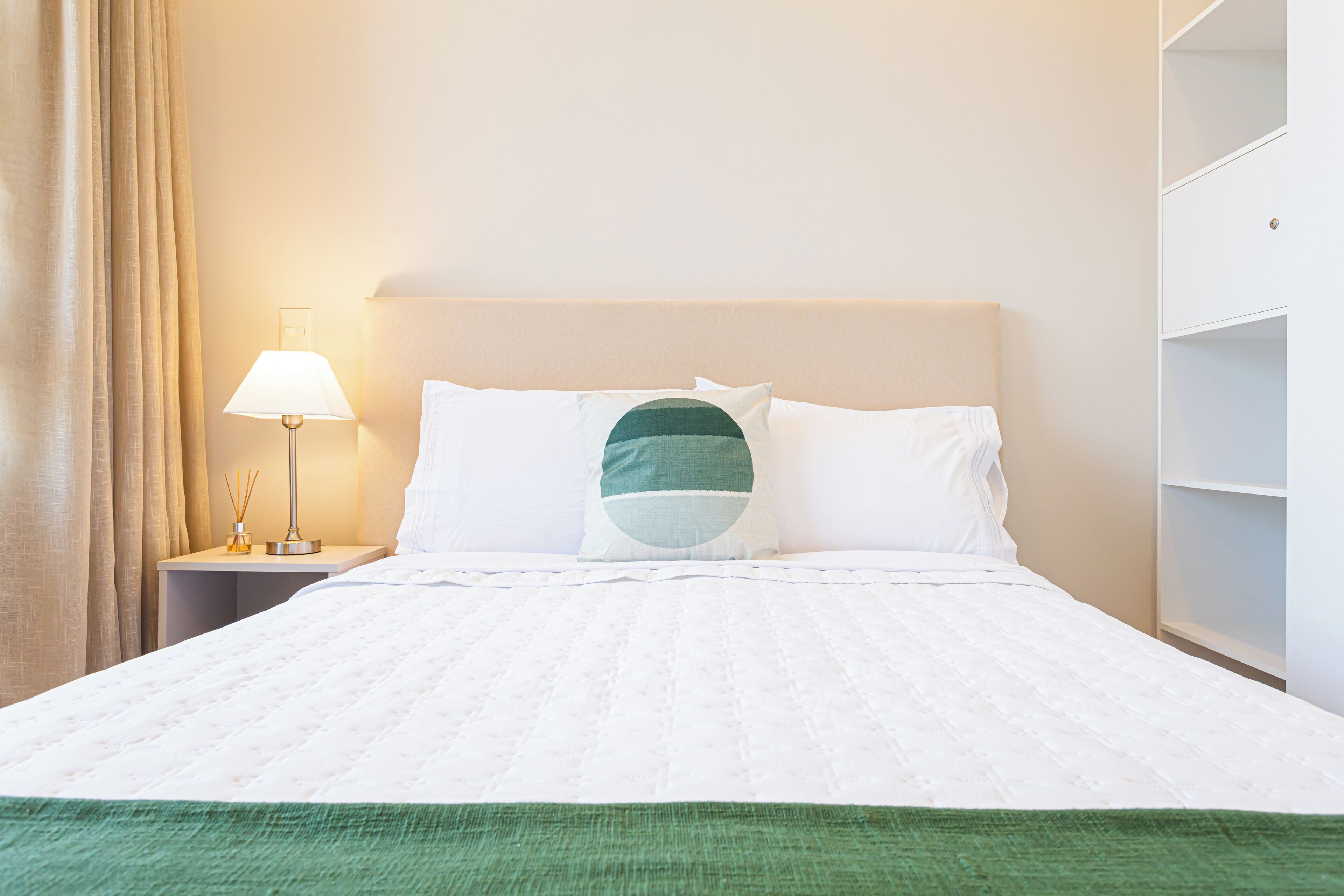A neatly made bed with white bedding and a green throw at the foot. There is a bedside lamp with a white shade and a small table with a lamp and a candle holder.