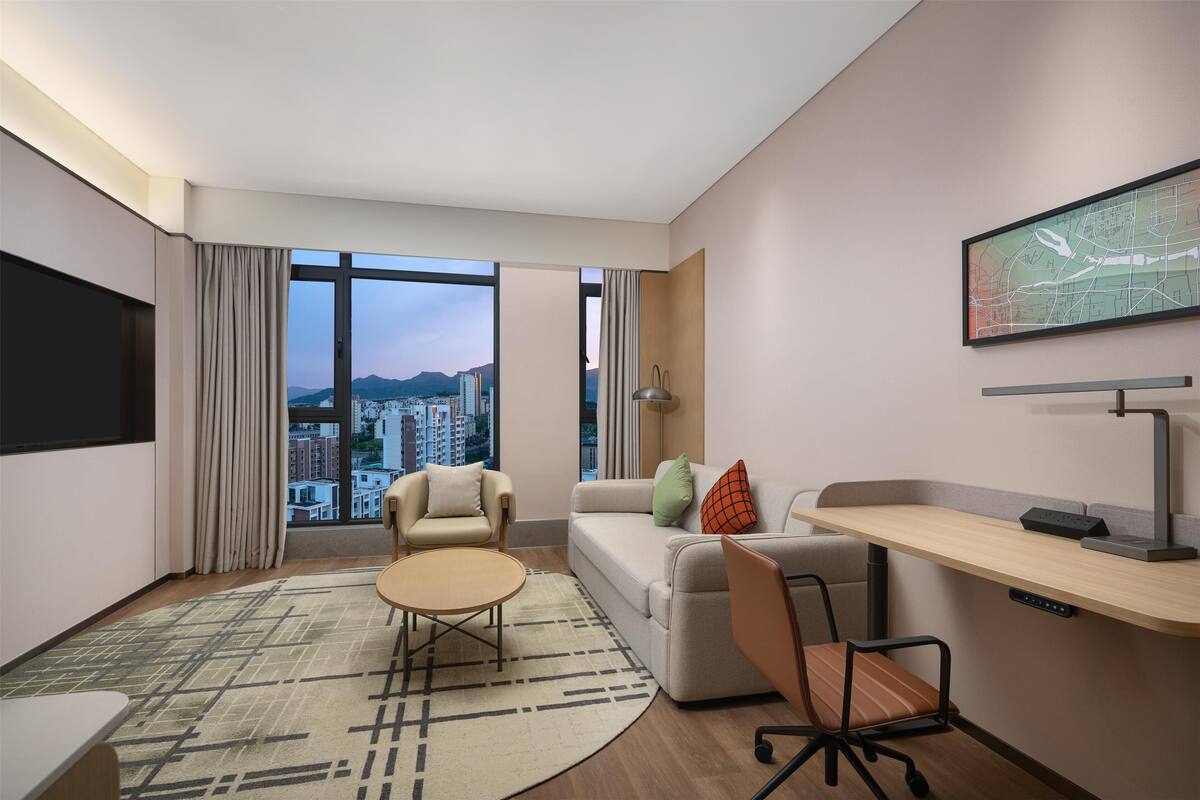 A modern hotel room with a large window, a sofa, a desk, and a chair.