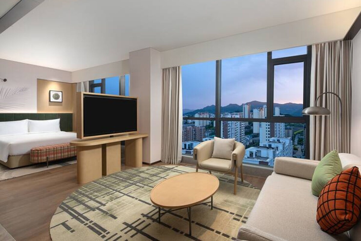 A hotel room with a bed, a TV, a sofa, a coffee table, and a large window with a city view.