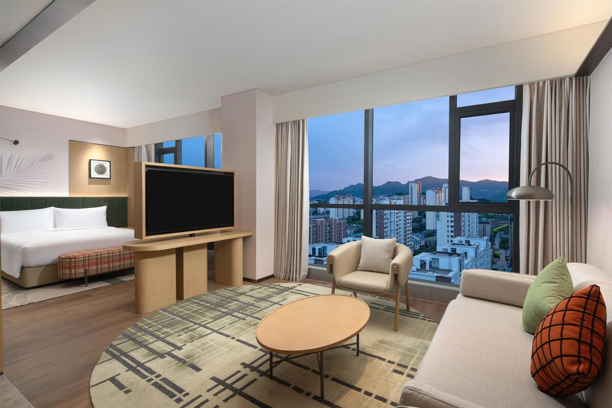 A modern hotel room with a bed, a sofa, a TV, and a large window offering a city view.