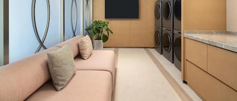 A modern laundry room with a flat-screen TV, stacked washer and dryer units, a beige sofa with cushions, and a potted plant.