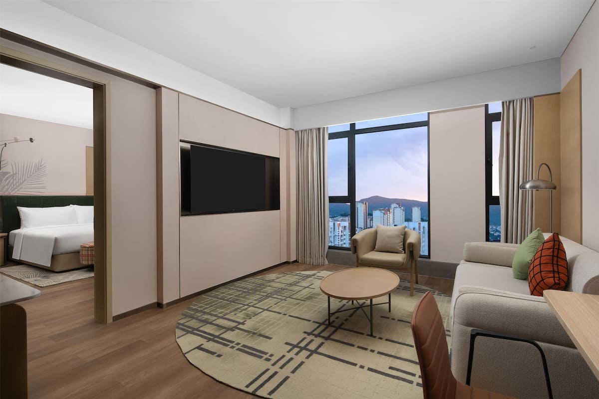 A modern hotel room with a bed, a flat-screen TV, a sofa, a coffee table, and a view of the city.