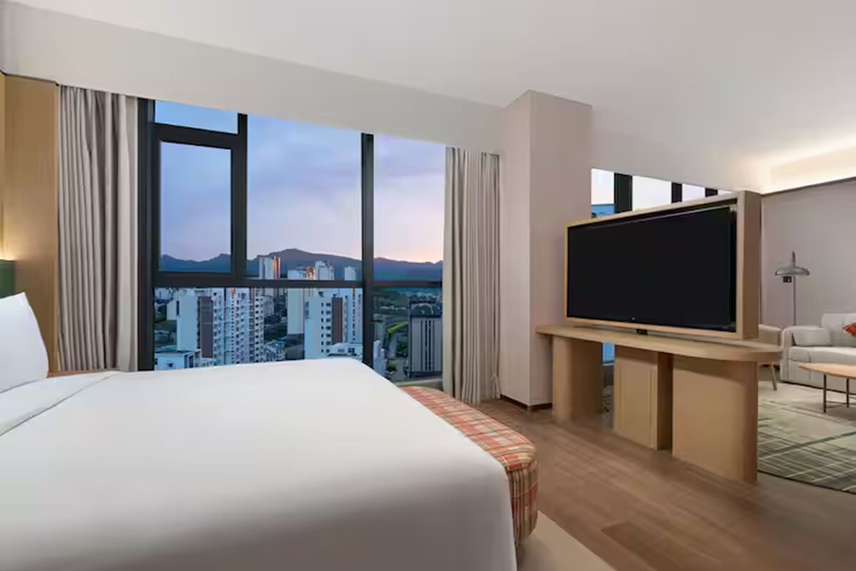Habitación de hotel con una cama grande, un televisor de pantalla plana y vistas a la ciudad a través de amplias ventanas.