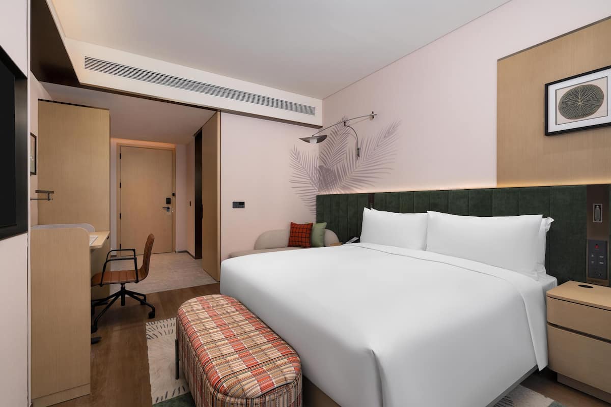 A modern hotel room with a large bed, a desk, and a chair. There is a bench at the foot of the bed, a nightstand, and a wall-mounted light fixture.