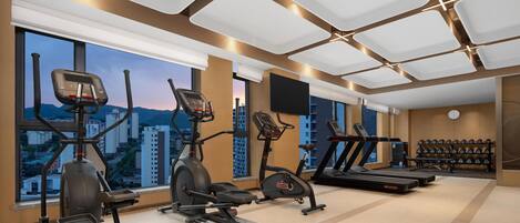 A modern gym with cardio machines, dumbbells, and a large window with a city view.