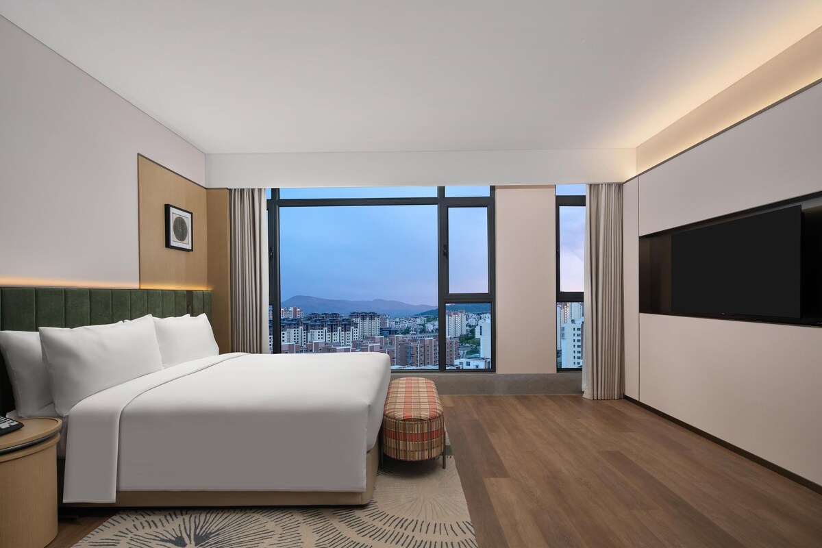 A modern hotel room with a large bed, a flat-screen TV, and a city view through large windows.