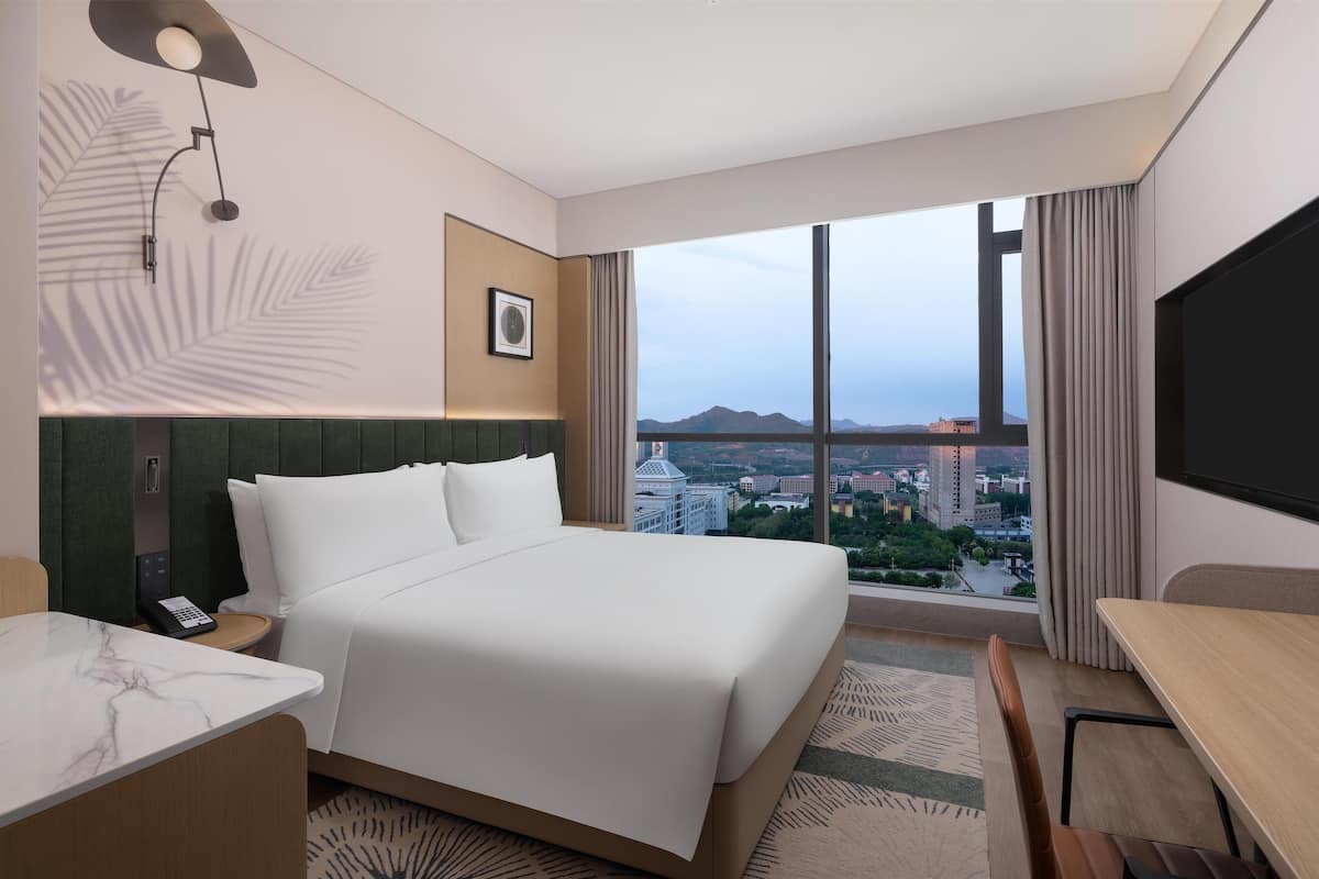 A hotel room with a large bed, a desk, a chair, a telephone, and a large window offering a city view.