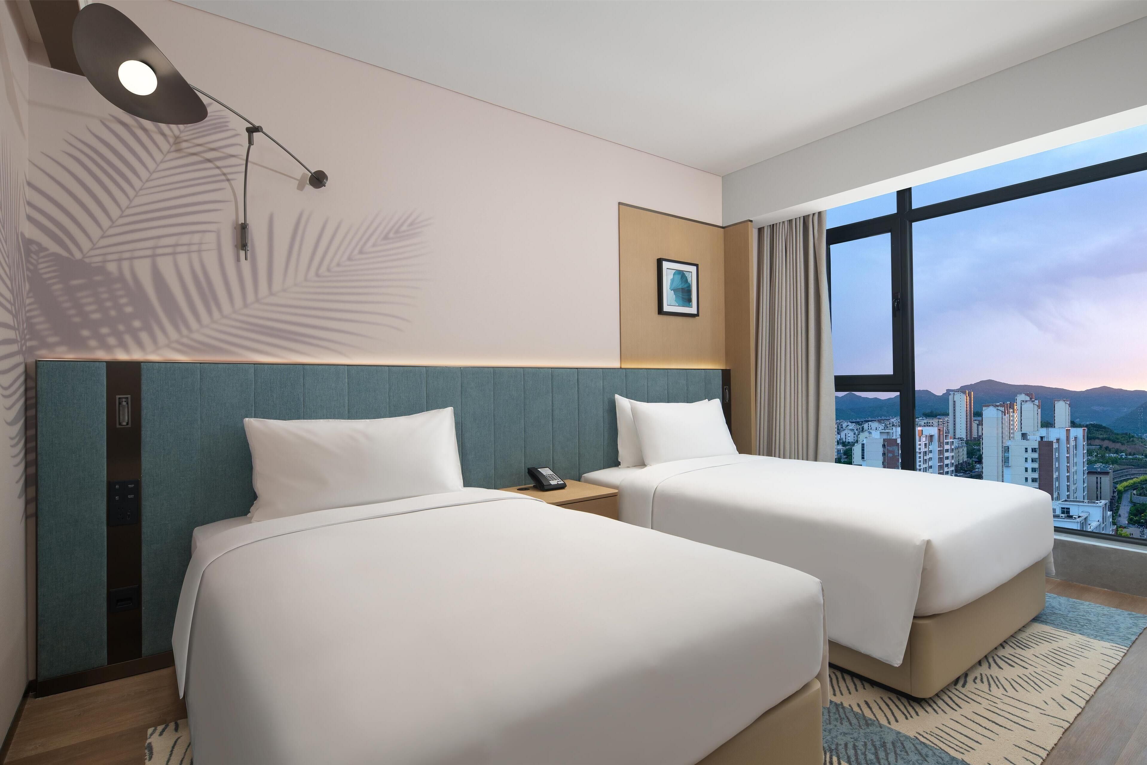 A hotel room with two beds, a large window with a city view, a wall-mounted lamp, and a small picture.