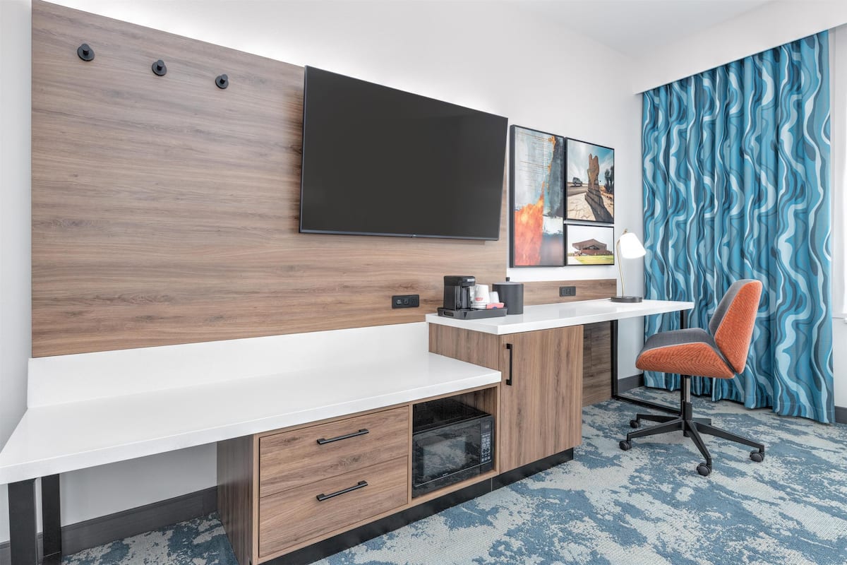 A modern hotel room with a flat-screen TV mounted on a wooden wall, a white desk with a coffee maker, a microwave, and a chair.