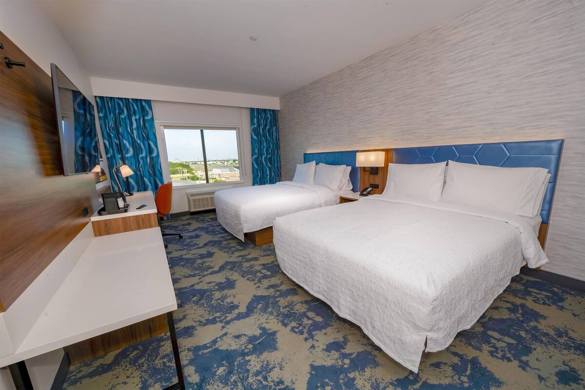 A hotel room with two beds, a desk, a mirror, and a window with a view.