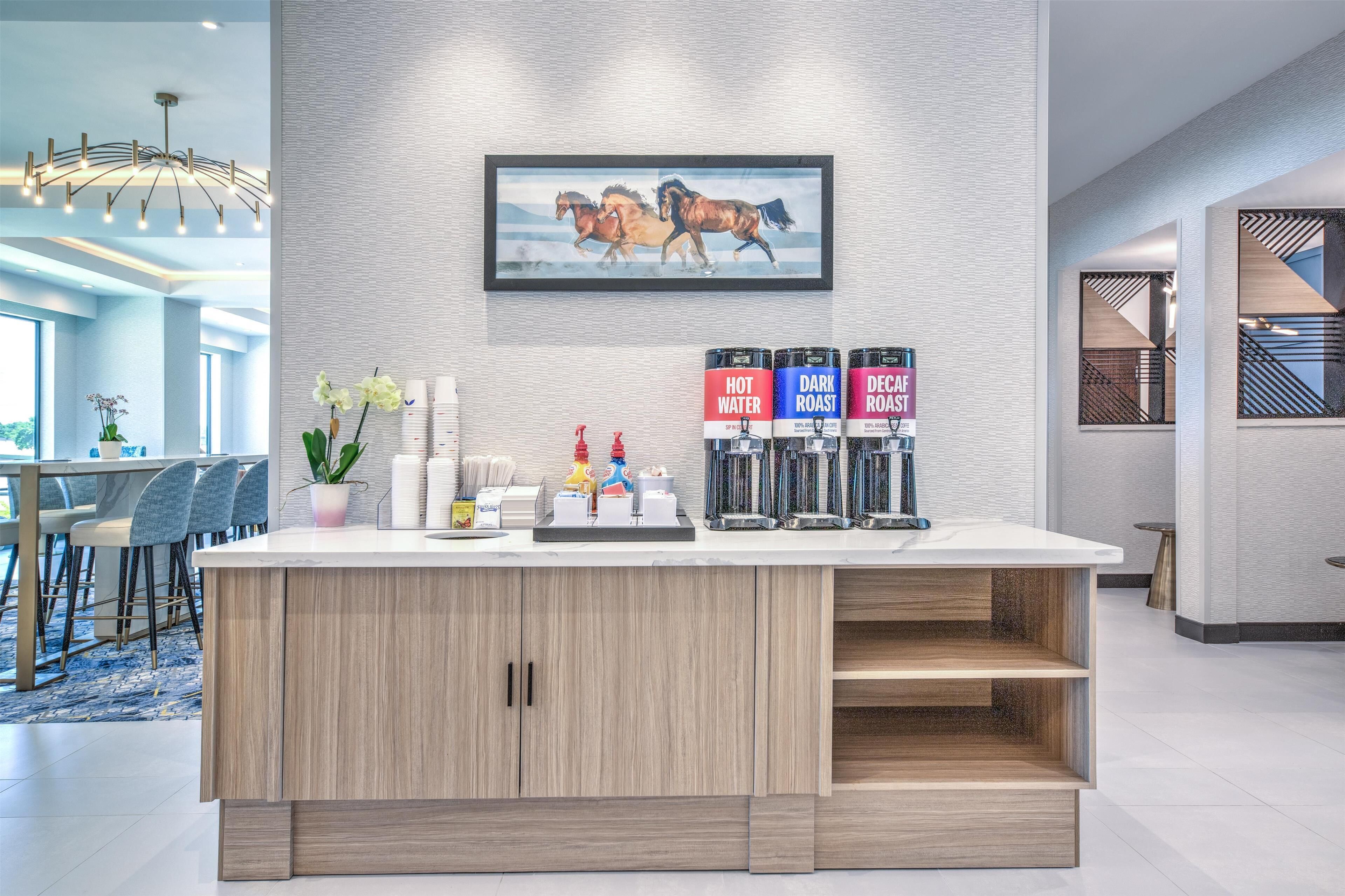 A modern kitchen area with a central island, a painting of horses on the wall, and a coffee station with labeled dispensers.