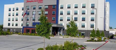 Um hotel Hampton Inn & Suites com fachada moderna, várias janelas e estacionamento com áreas ajardinadas.