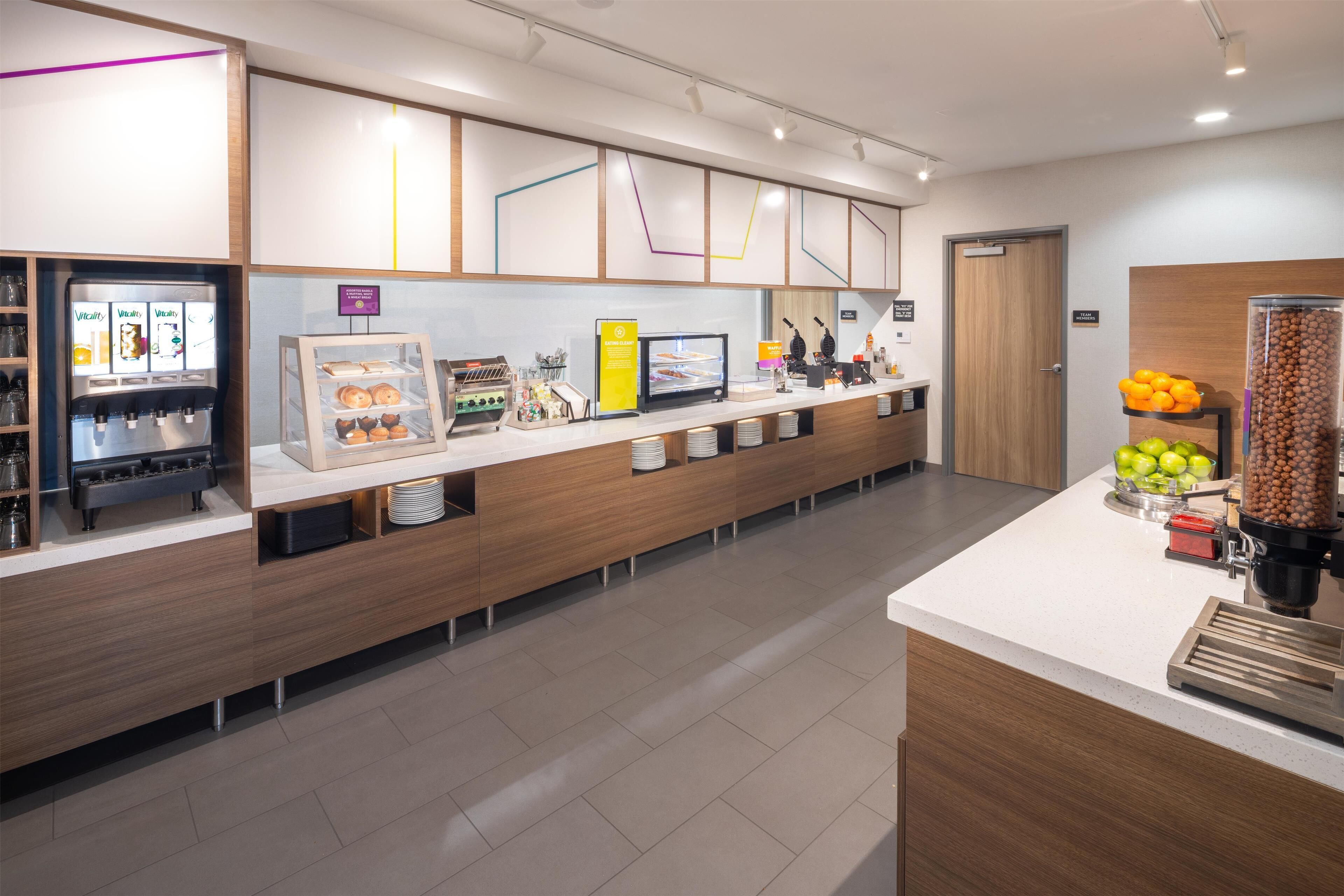 Ein moderner Selbstbedienungsbereich für das Frühstück mit einer Kaffeemaschine, einer Vitrine voller Gebäck und einer Obstschale.