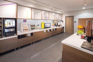 A modern self-service breakfast area with a coffee machine, a display case with pastries, and a fruit bowl. - Home2 Suites by Hilton Kalispell Whitefish (Kalispell)