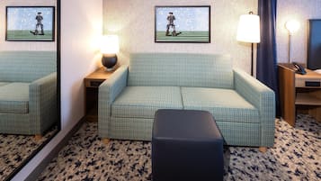 Une chambre d’hôtel avec un canapé à motifs verts, un pouf noir, une petite table avec une lampe et deux tableaux encadrés accrochés au mur.