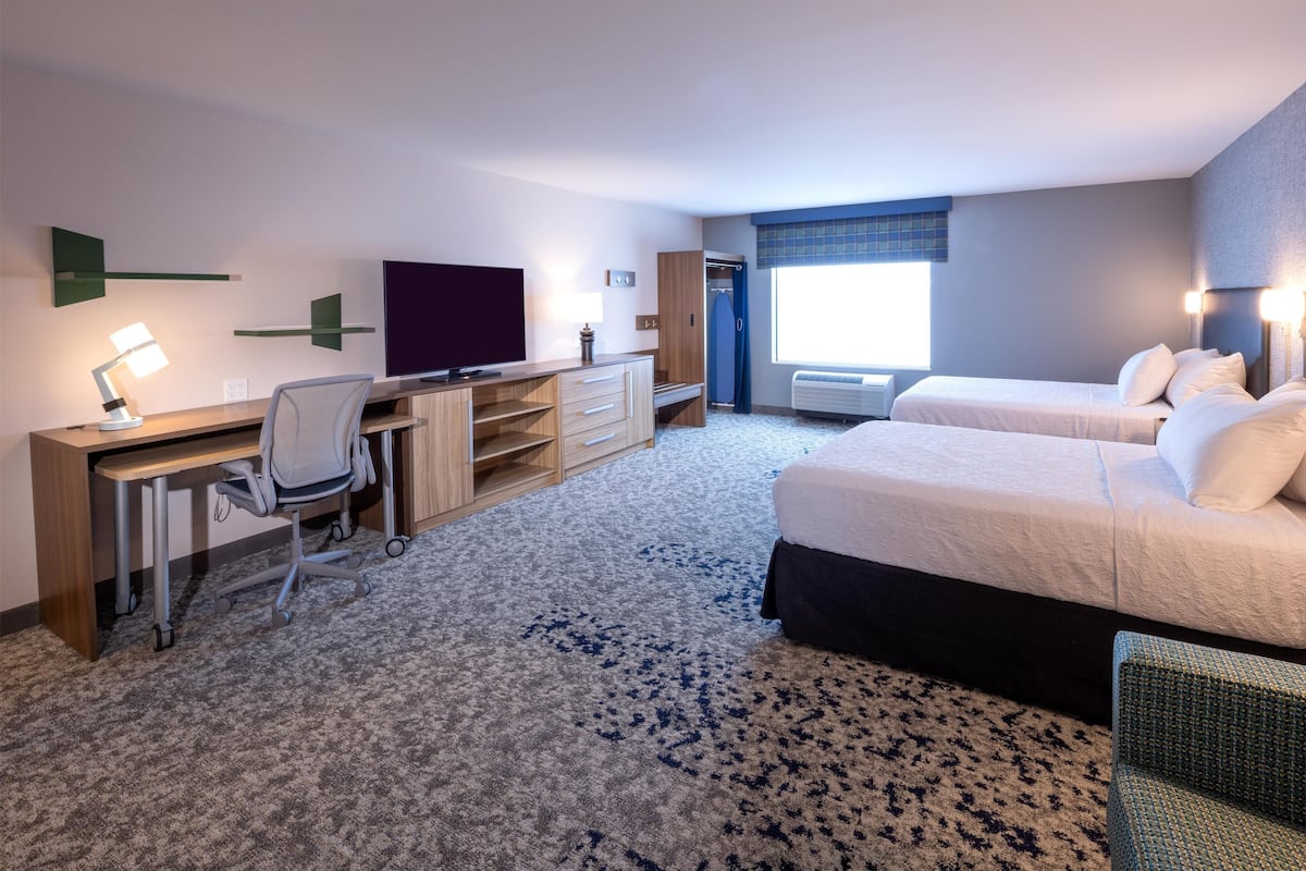 A hotel room with a desk, chair, two beds, a TV, and a window with curtains.