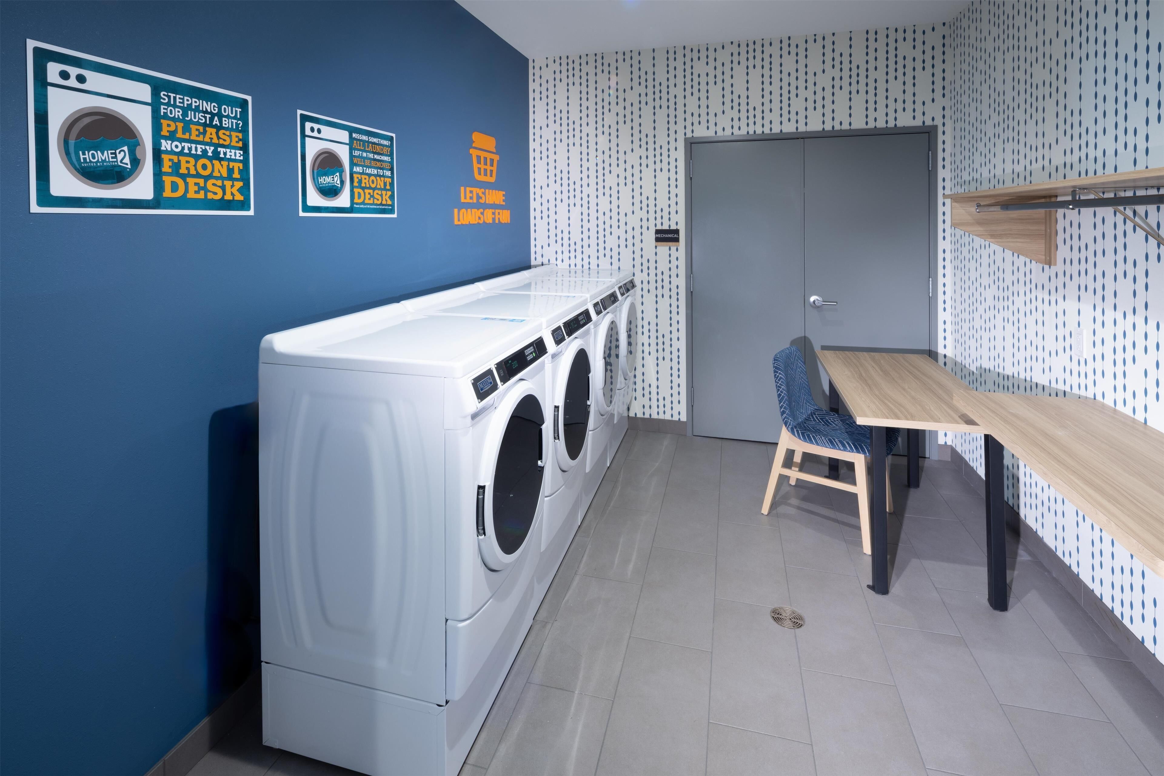 A laundry room with a washing machine, a drying rack, and a table with chairs. There are signs on the wall and a door with a handle.