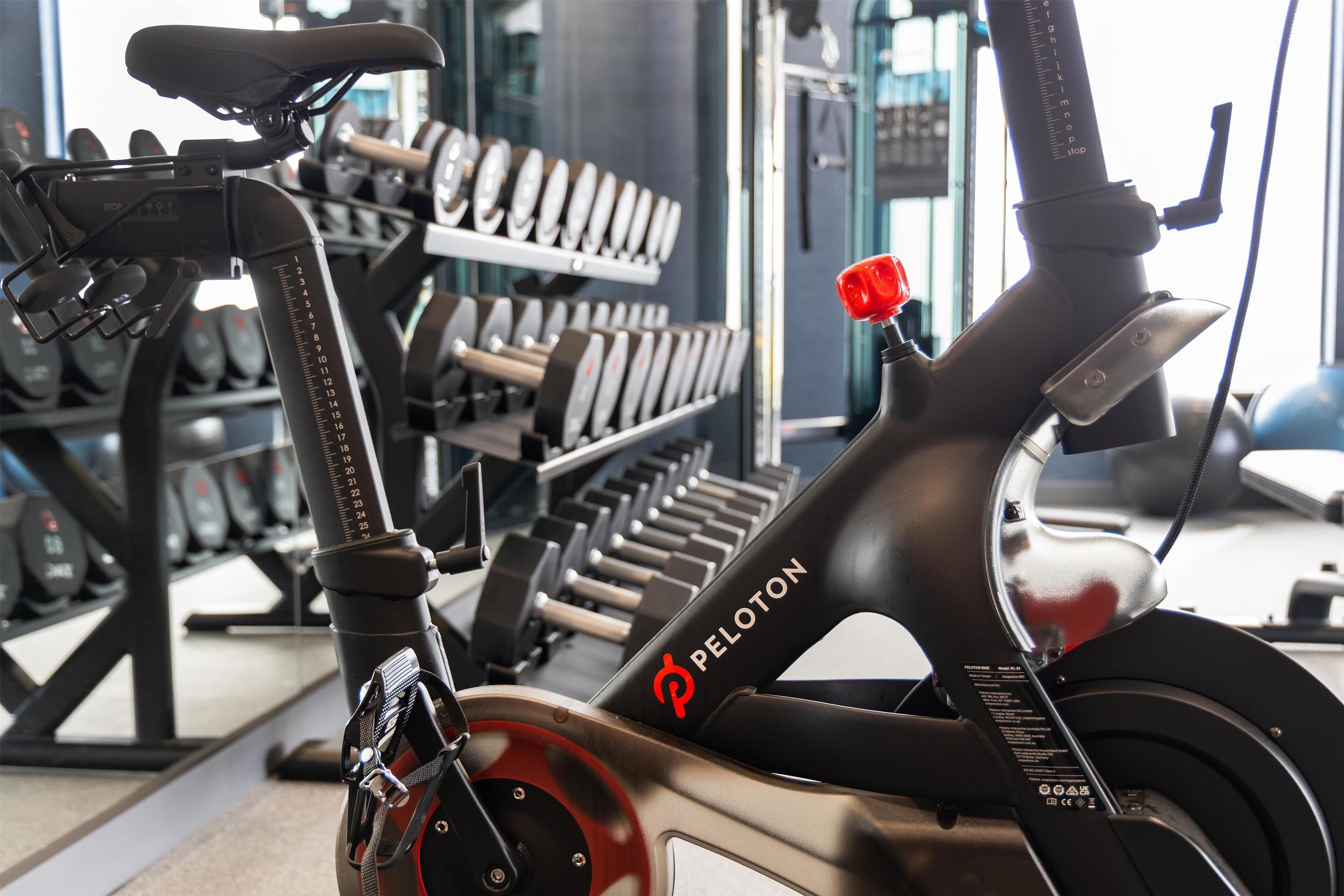 Un vélo d'appartement Peloton dans une salle de sport, avec des appareils de musculation en arrière-plan.