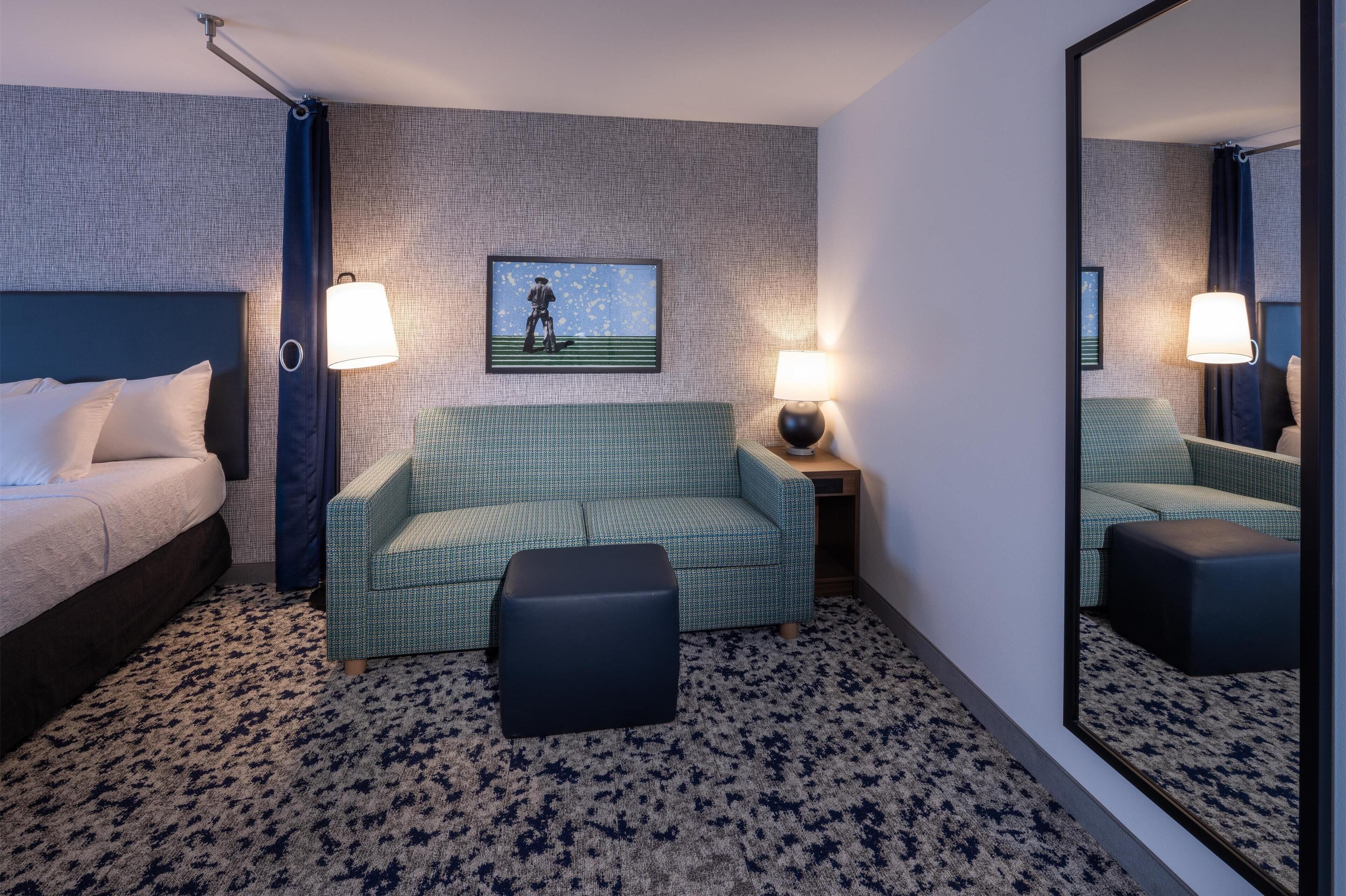 Une chambre d’hôtel avec un lit, un canapé vert, un pouf assorti, une table de chevet avec une lampe et un tableau encadré au mur.