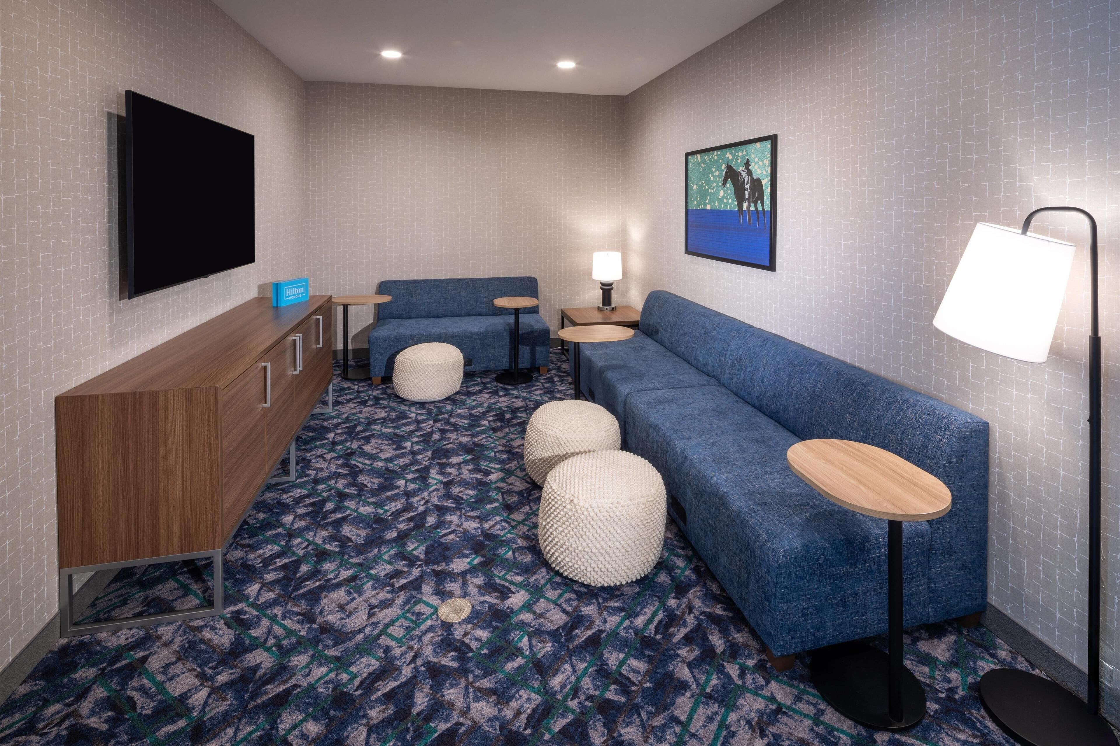 A modern hotel room with a blue sofa, a wooden cabinet, a flat-screen TV, a lamp, and a patterned carpet.