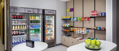 A modern snack bar with refrigerated drinks and snacks, a sign stating 'HOME IS WHERE THE SNACKS ARE', and a table with green apples.