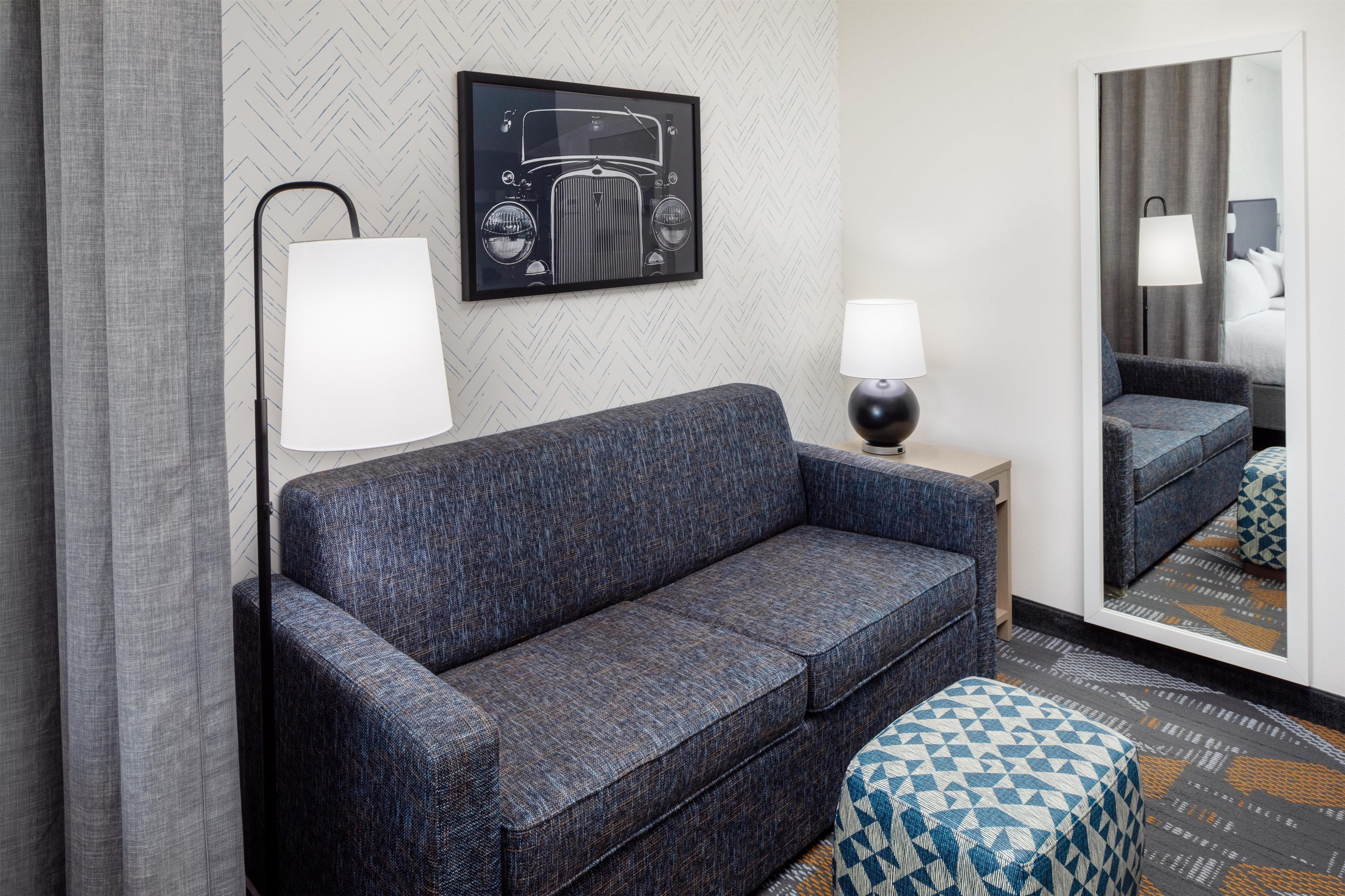 Habitación de hotel con un sofá azul oscuro, una mesita de noche con lámpara, un reposapiés estampado y un espejo grande que refleja la habitación.