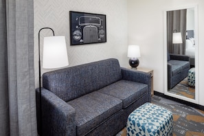 A hotel room with a dark blue sofa, a side table with a lamp, a patterned ottoman, and a large mirror reflecting the room. - Home2 Suites By Hilton St. Joseph (St. Joseph)