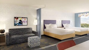 A hotel room with a sofa, two beds, a patterned ottoman, a wall art piece, and a window with a view. - Home2 Suites By Hilton St. Joseph (St. Joseph)