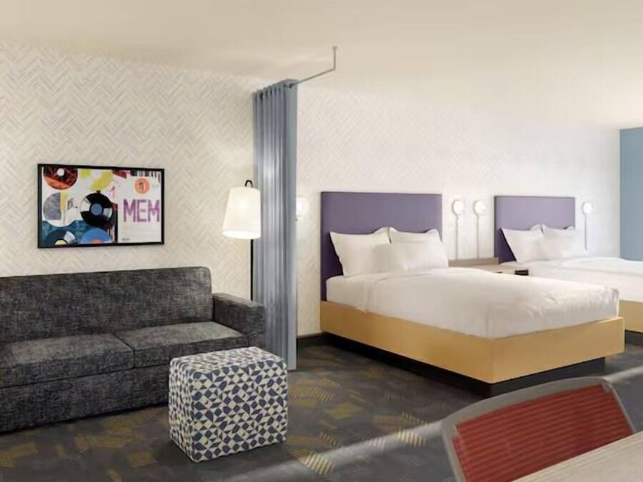 A hotel room with a sofa, two beds, a patterned ottoman, a wall art piece, and a window with a view.