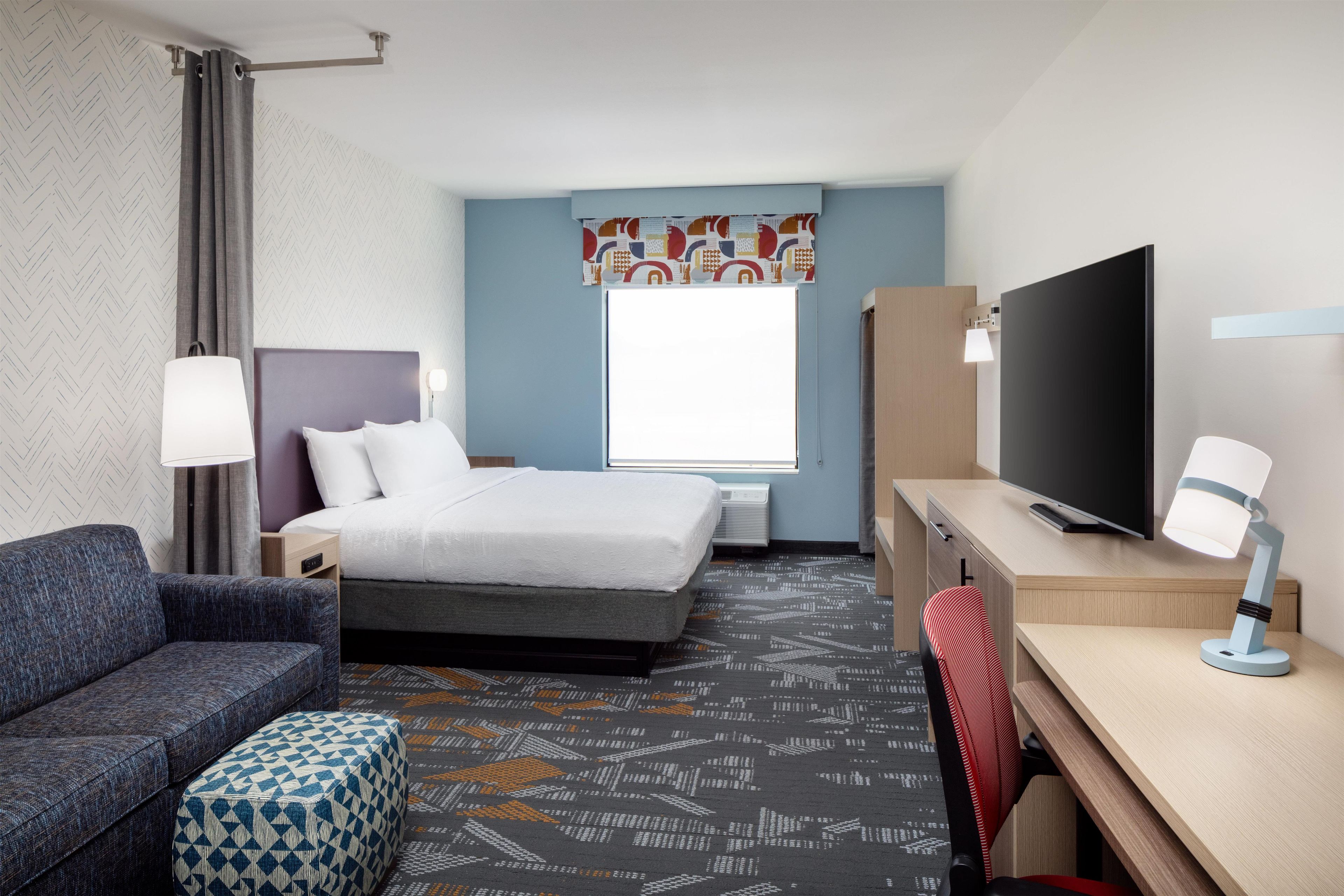 A hotel room with a bed, a desk, a TV, a chair, and a patterned carpet.