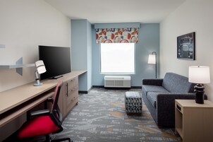 A hotel room with a desk, chair, television, sofa, ottoman, and a window with a patterned curtain.