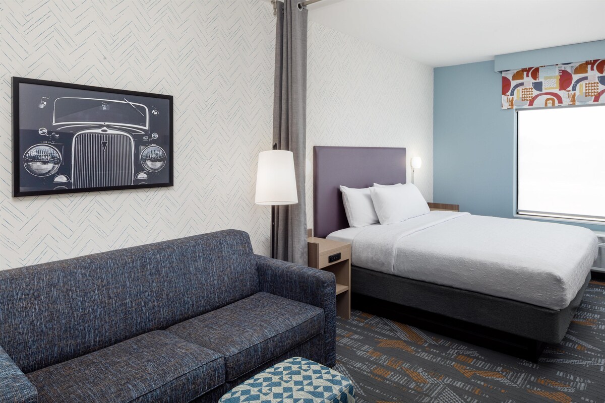 A hotel room with a bed, a sofa, a nightstand, a lamp, and a wall art piece.