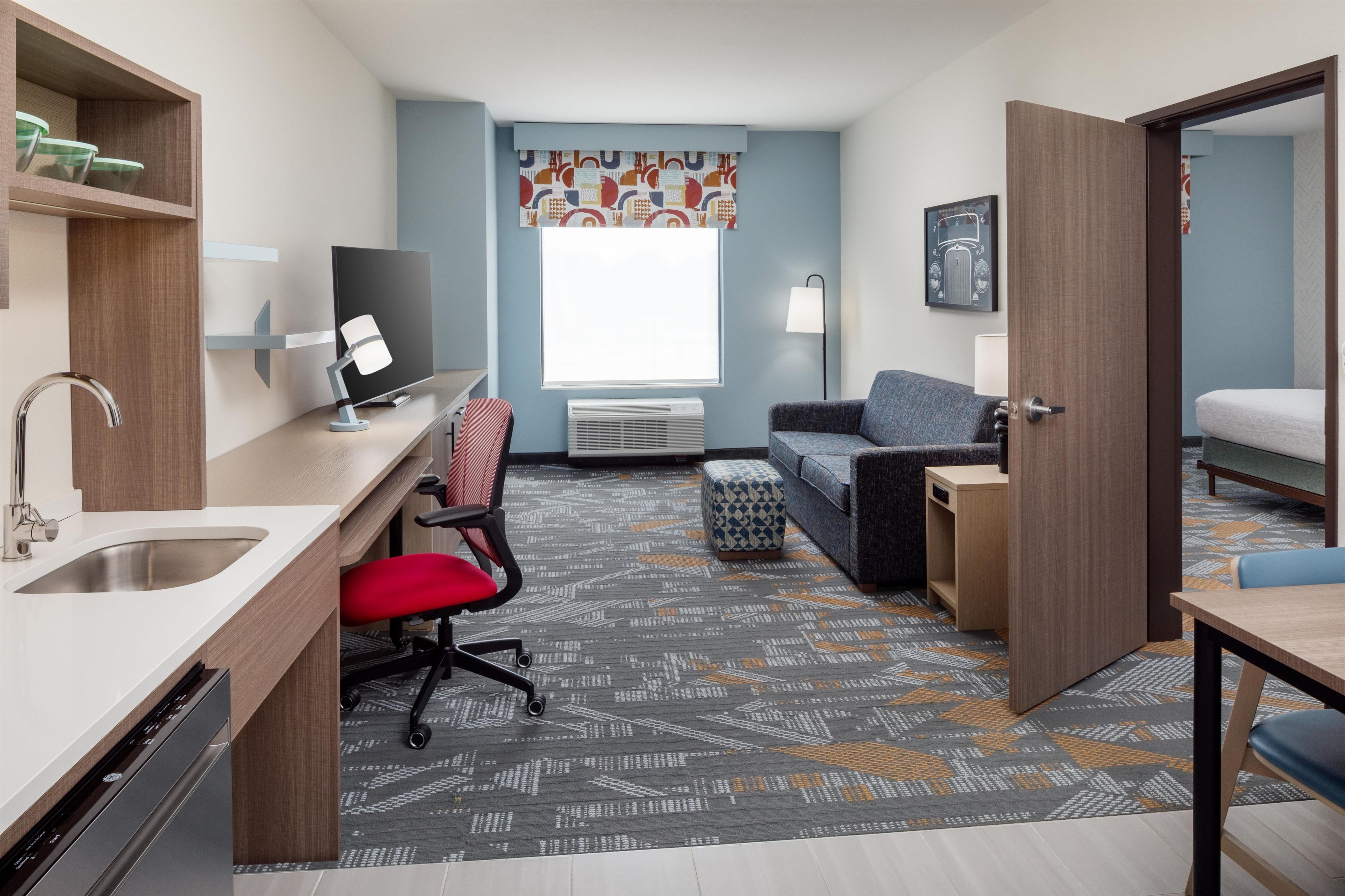 A hotel room with a kitchenette, a desk with a computer, a chair, a sofa, and a bed.