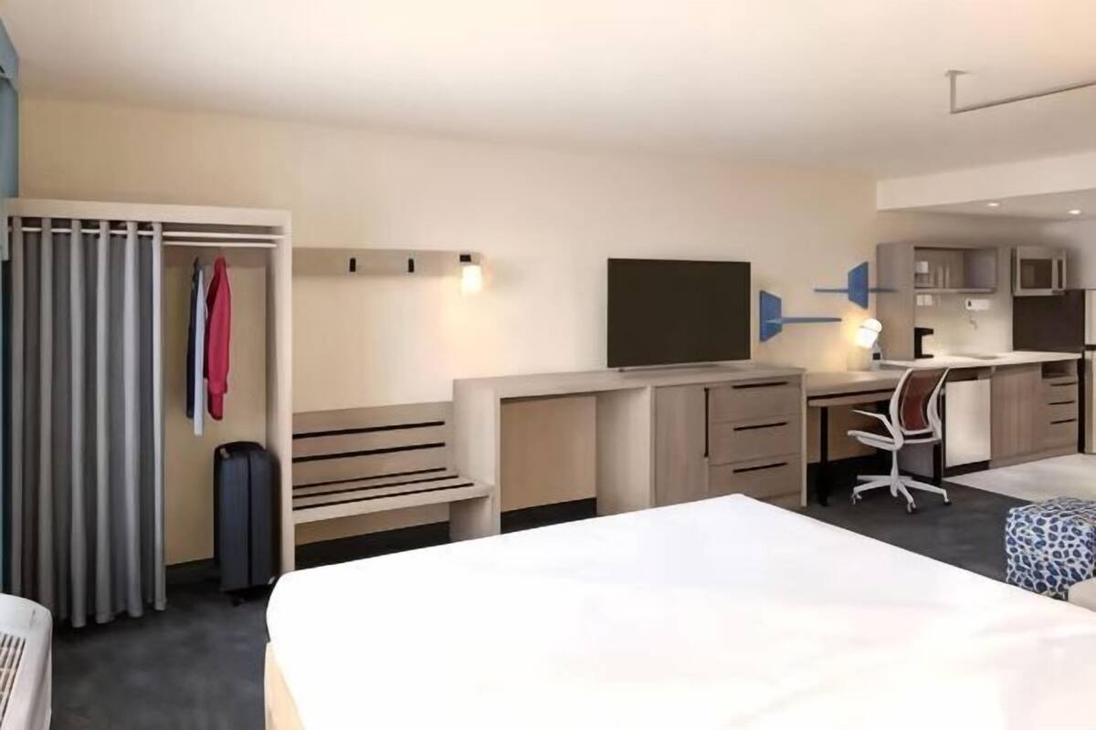 A hotel room with a bed, desk, chair, TV, wardrobe, and a small kitchenette.