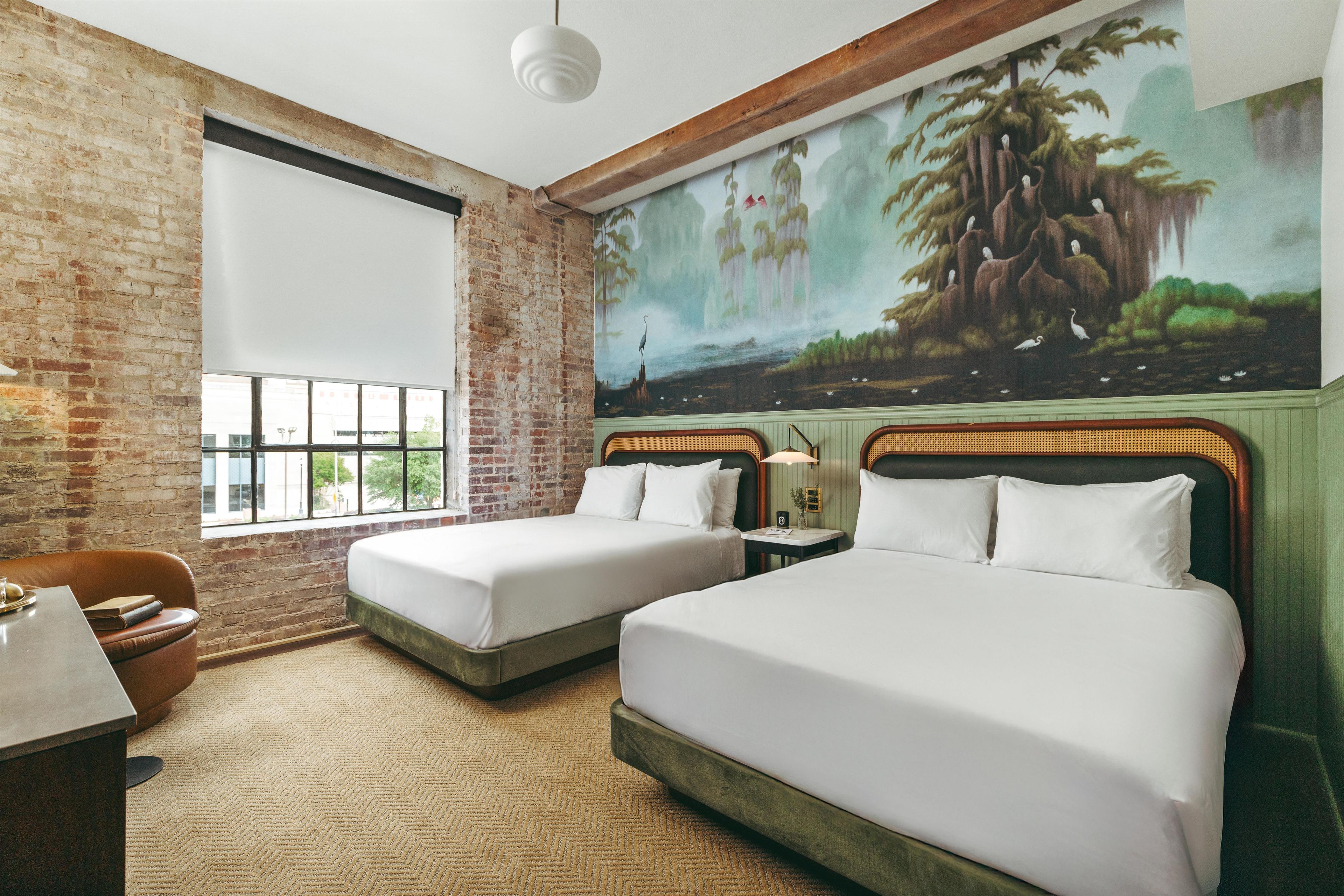 Una habitación de hotel con dos camas, un ventanal grande, una pared de ladrillo y un mural.